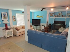 A living room with a blue sofa, a television, a fireplace, and various wall decorations including a fish and a surfboard. - Canal Front Home on sought out street - Unique, Cozy and Ready for YOU! (Ocean Isle Beach)