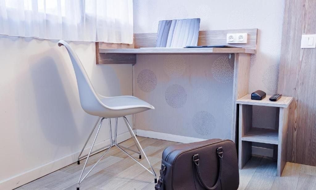 A room with a white chair, a wooden desk, a small shelf with a remote control, and a suitcase.