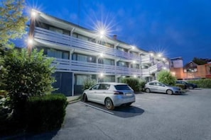 Un hotel de varios pisos con estacionamiento, varios automóviles aparcados y una fachada bien iluminada.