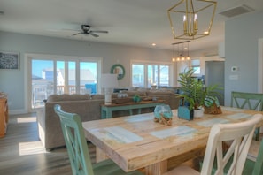 A spacious living room with a wooden dining table, green chairs, a sofa, and a ceiling fan. - Boozy Barradcuda: Ocean View, Pool, Game Room. Walk to Beach, Sleeps 16 (Oak Island)