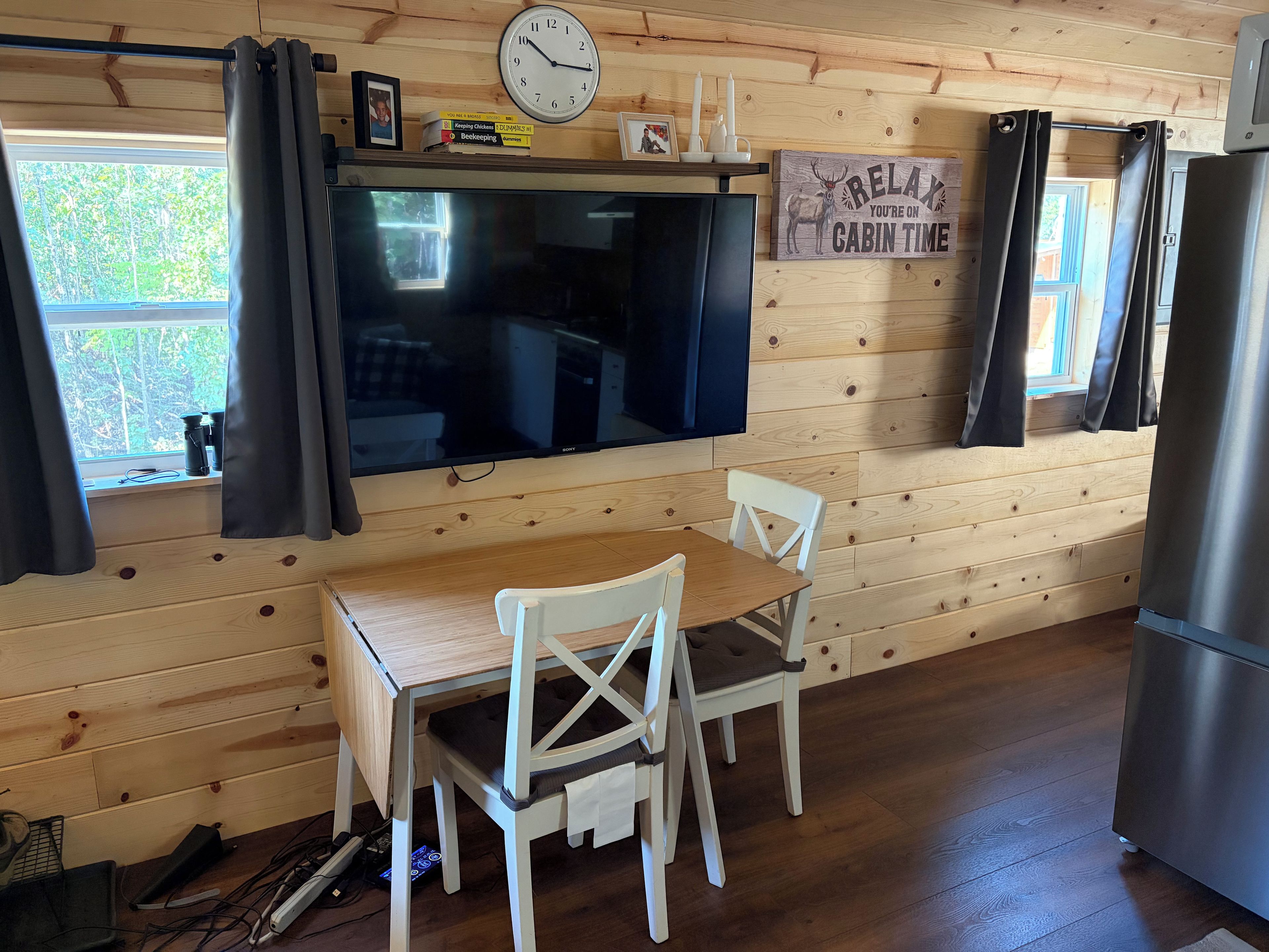 Interior d'una cabana amb paret de fusta, televisió de pantalla plana, taula de dinar amb cadires, nevera i finestra amb cortines.