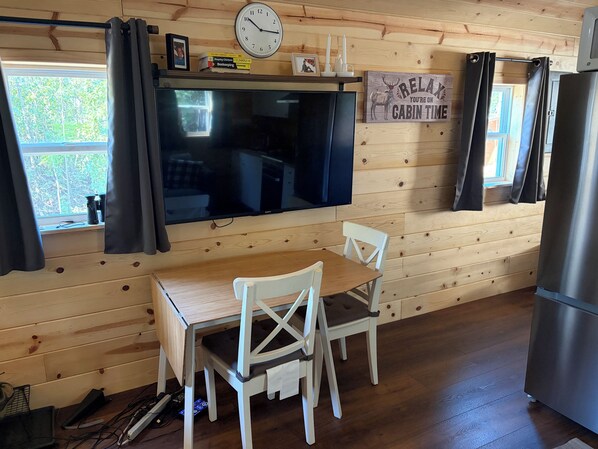 A cabin interior with a wooden wall, a flat-screen TV, a dining table with chairs, a refrigerator, and a window with curtains.