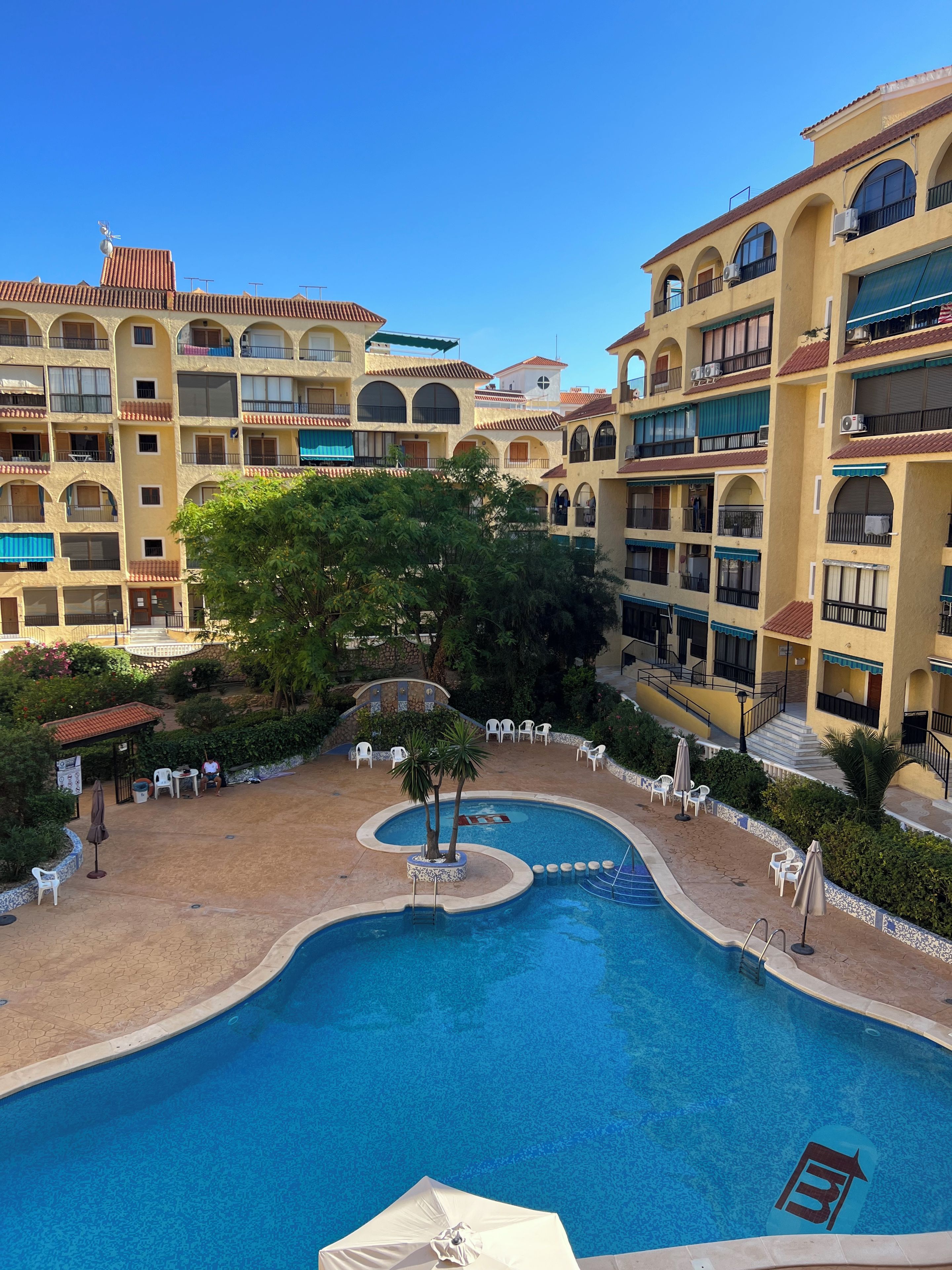 A hotel courtyard with a swimming pool, outdoor seating, and multi-story buildings with balconies.