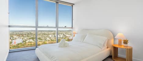 Un dormitorio con un ventanal grande, una cama, dos lámparas de noche y vistas a un paisaje urbano.