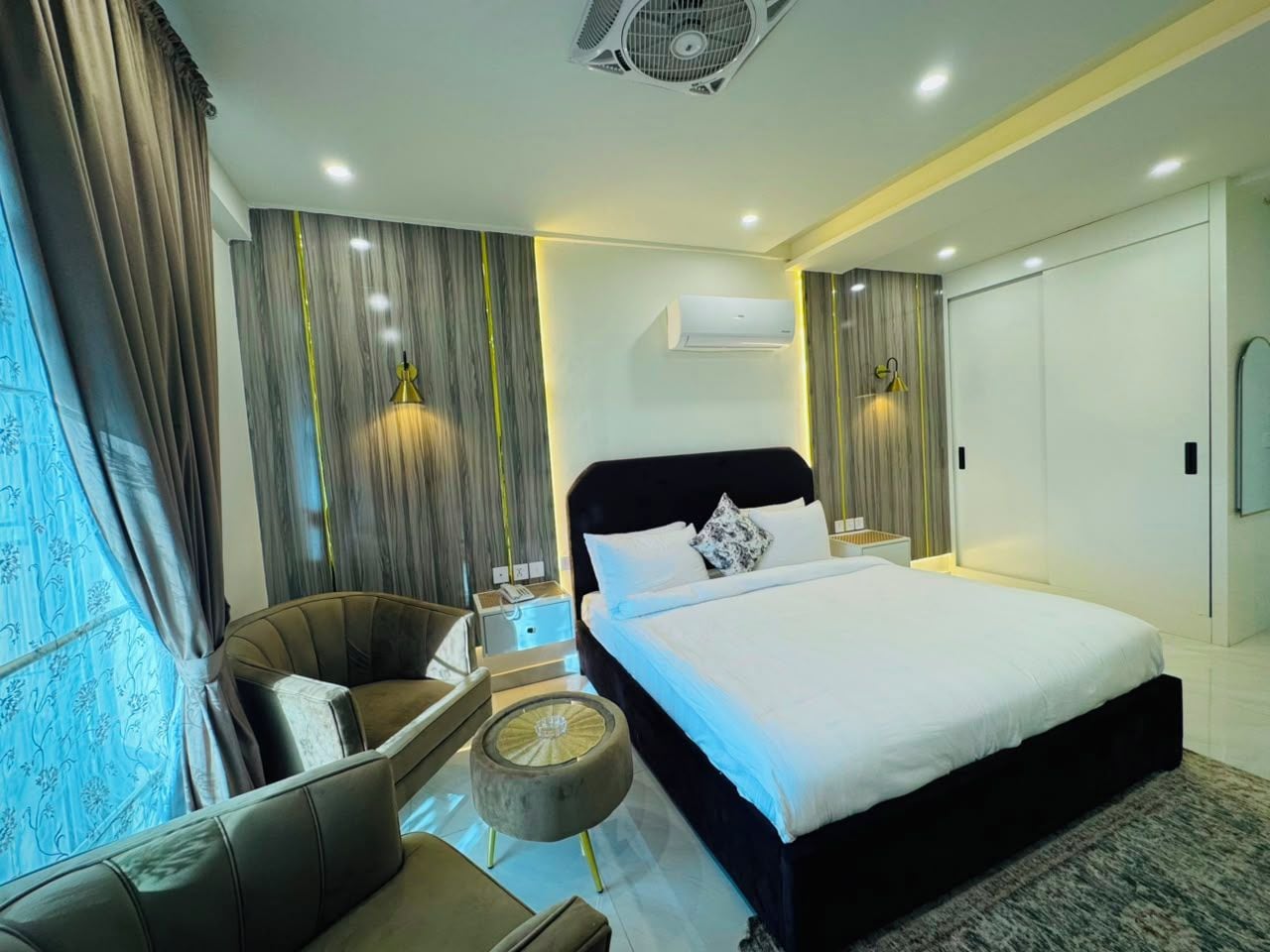 A modern hotel room with a large bed, a sitting area with a chair and a small table, and a large window with curtains.