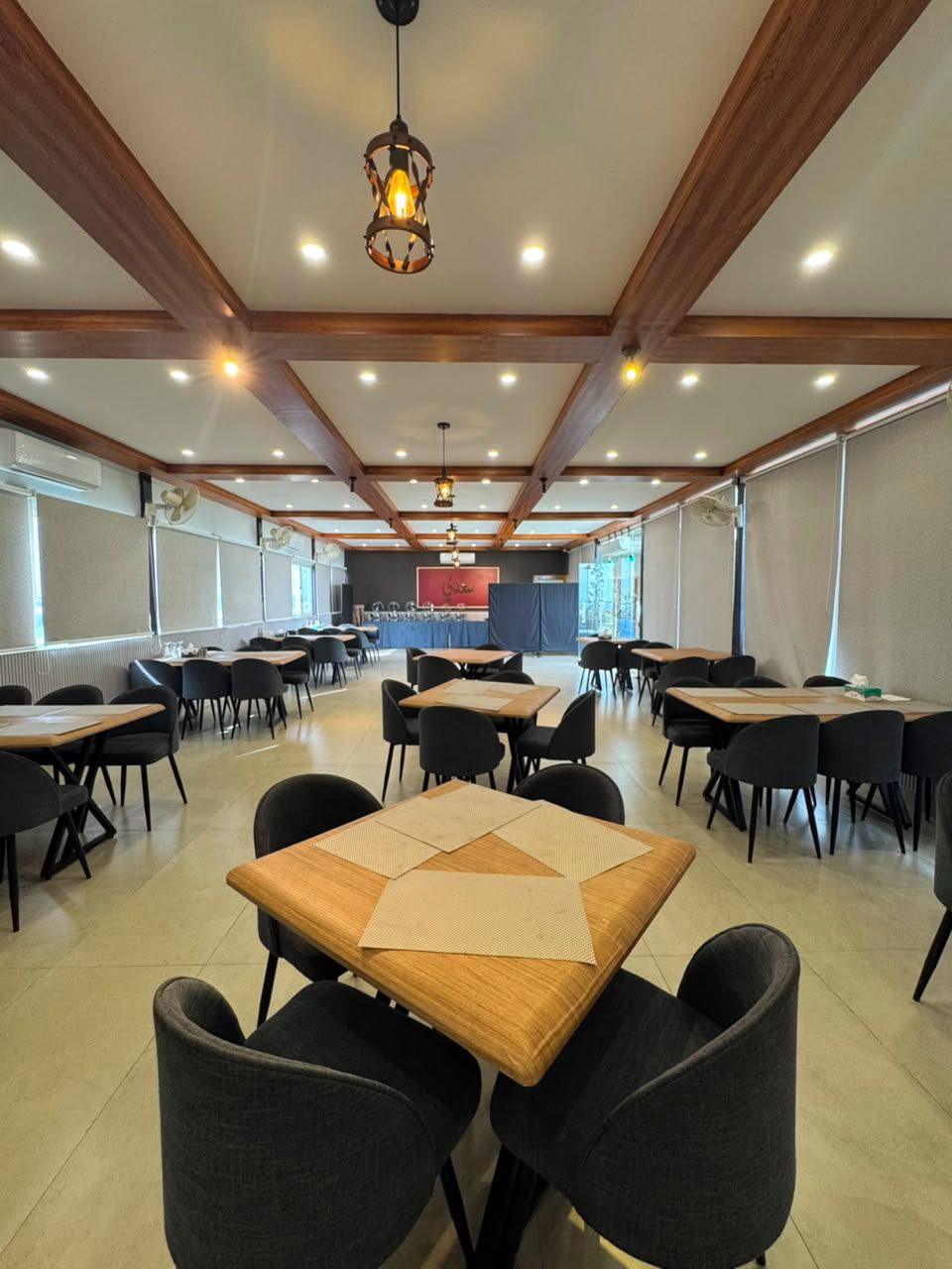 A spacious indoor dining area with multiple tables and chairs, wooden beams on the ceiling, and a central light fixture.