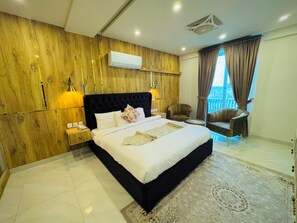 Habitación de hotel con una cama grande, un revestimiento de madera en la pared, un ventilador de techo, una mesita de noche con un teléfono y vistas a la ciudad a través de la ventana.