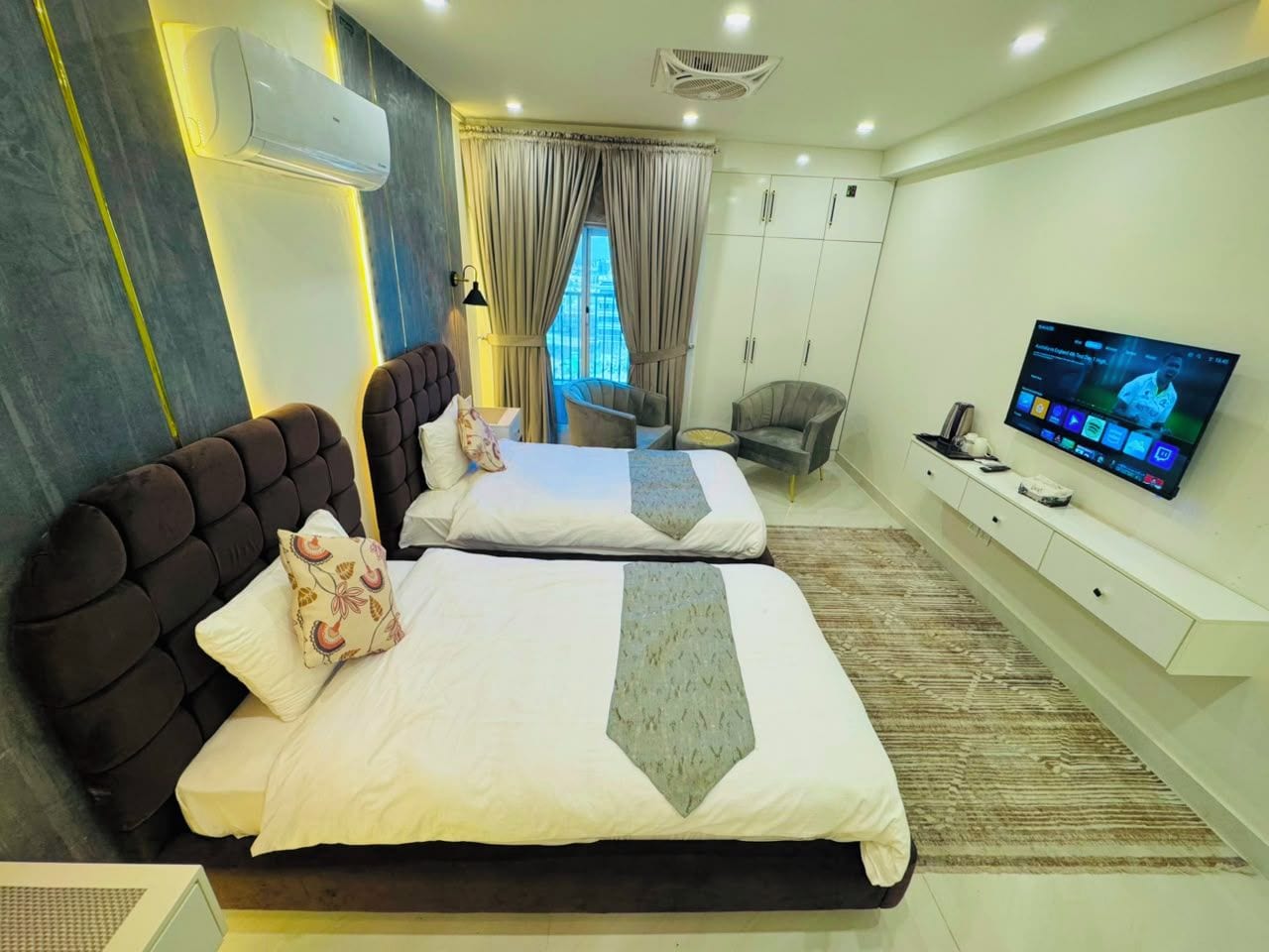 Quarto de hotel com duas camas, televisão, guarda-roupa e janela com cortinas.