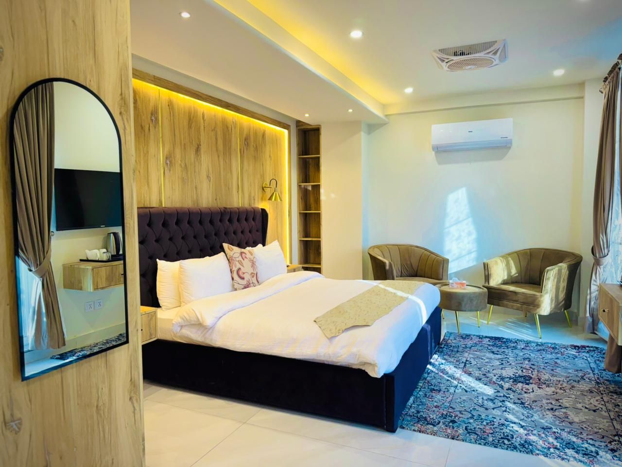 A modern hotel room with a large bed, a bedside table, a chair, a small table, and a mirror.