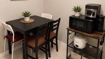 A dining area with a black table, two chairs, a framed plant, a microwave, and a rice cooker on a shelf.