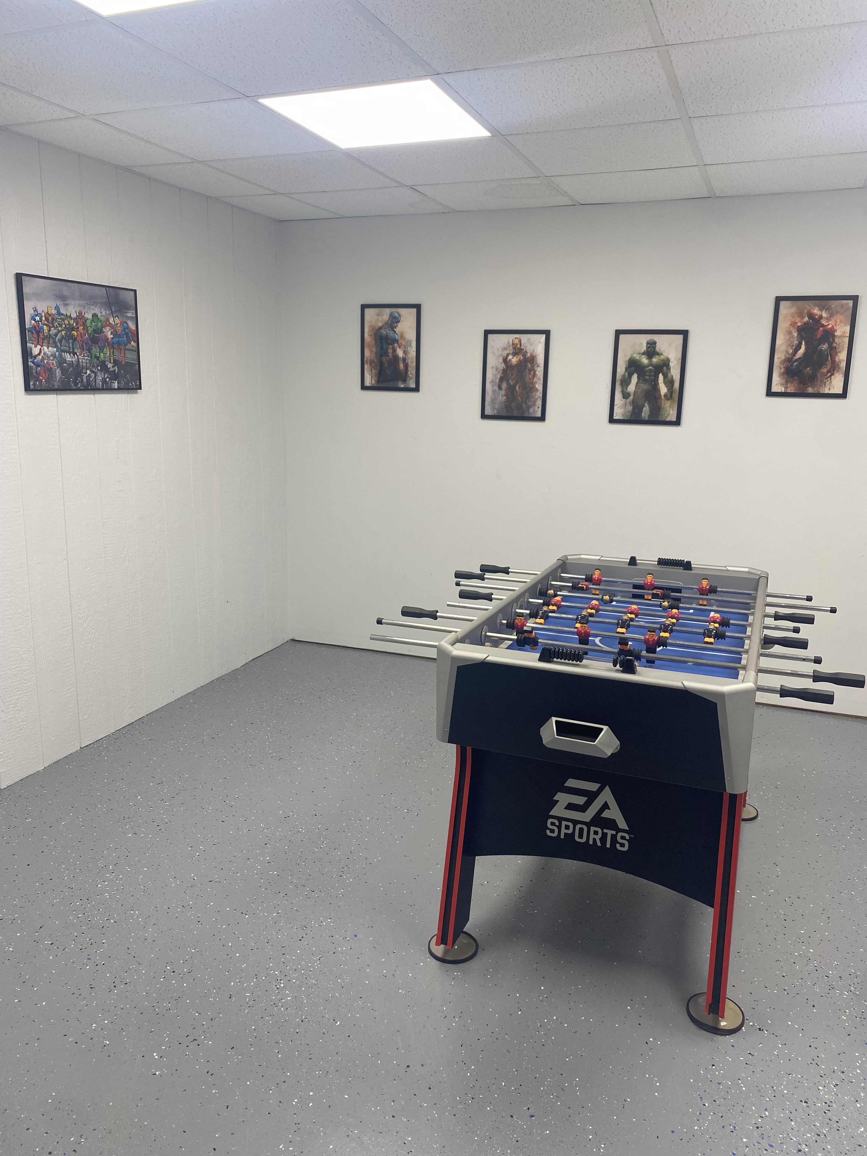 Une table de baby-foot dans une pièce aux murs blancs et au sol gris, avec quatre tableaux encadrés accrochés au mur.