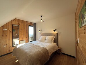 Ein Schlafzimmer mit einem Bett, einem Nachttisch, einer Holzbalkendecke und Blick durch eine offene Tür.