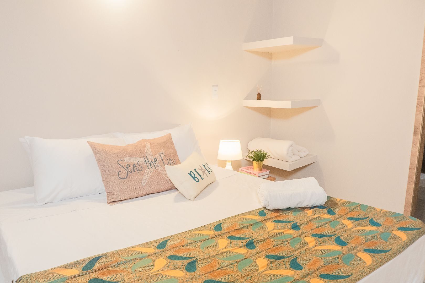 Ett hotellrum med en säng, kuddar, ett sängbord med en lampa, en liten växt och en handduk på en hylla.