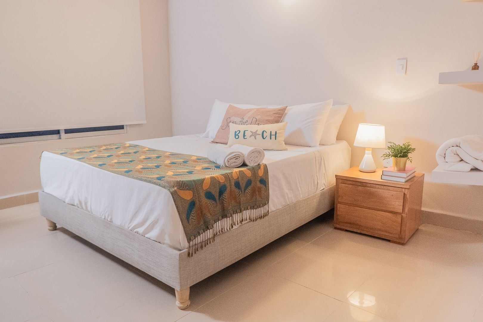 Una cama bien hecha con una colcha estampada, una lámpara de noche y una planta pequeña sobre una mesita de noche de madera.