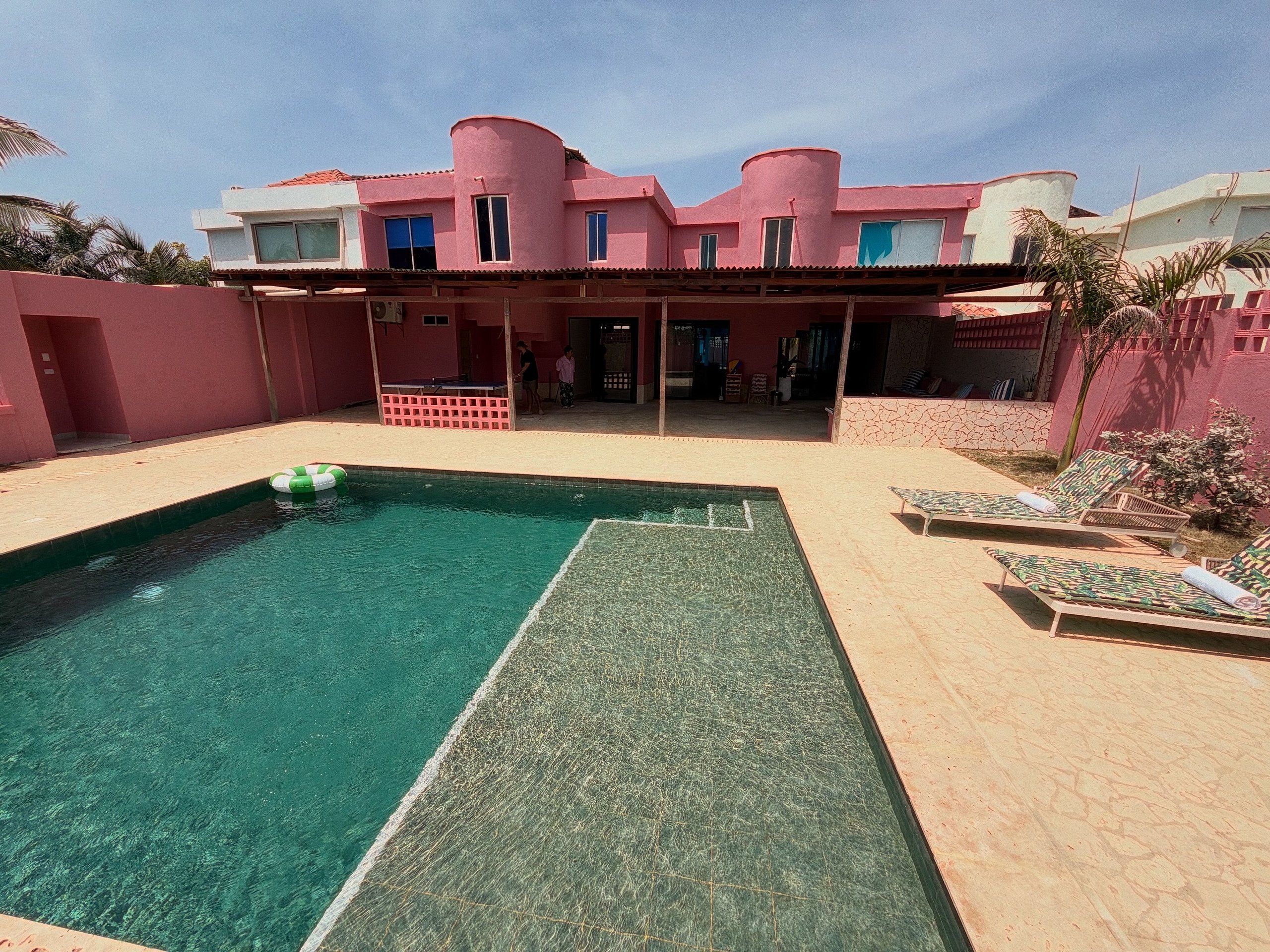 Piscina com deck verde, cadeiras de descanso e um edifício com fachada rosa.