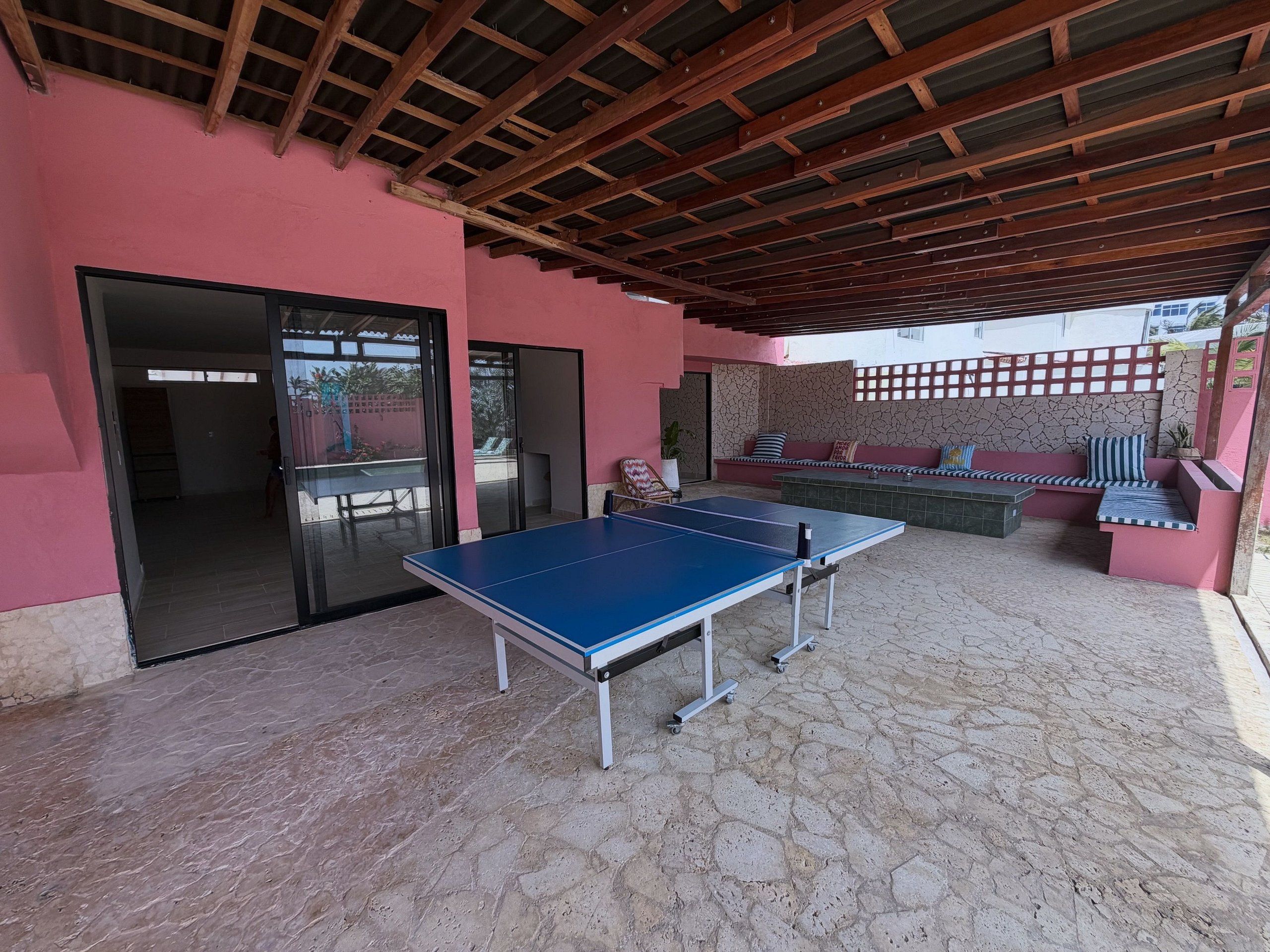 An outdoor area with a ping pong table, a seating area with cushions, and a covered space with a wooden ceiling.