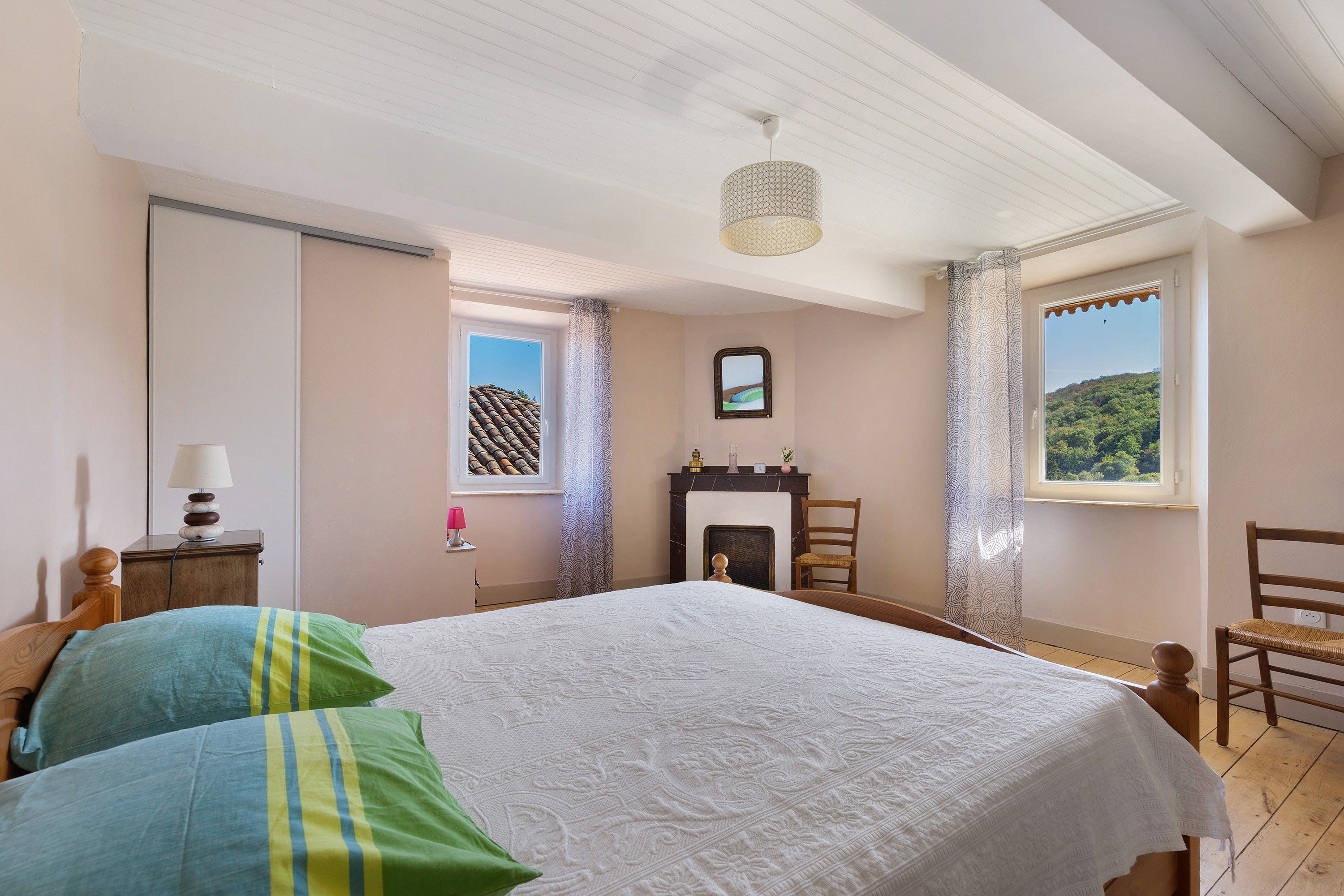 A bedroom with a bed, a nightstand, a lamp, a chair, a fireplace, and a window with a view of a hill.
