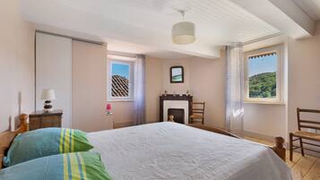 A bedroom with a bed, a nightstand, a lamp, a chair, a fireplace, and a window with a view of a hill.
