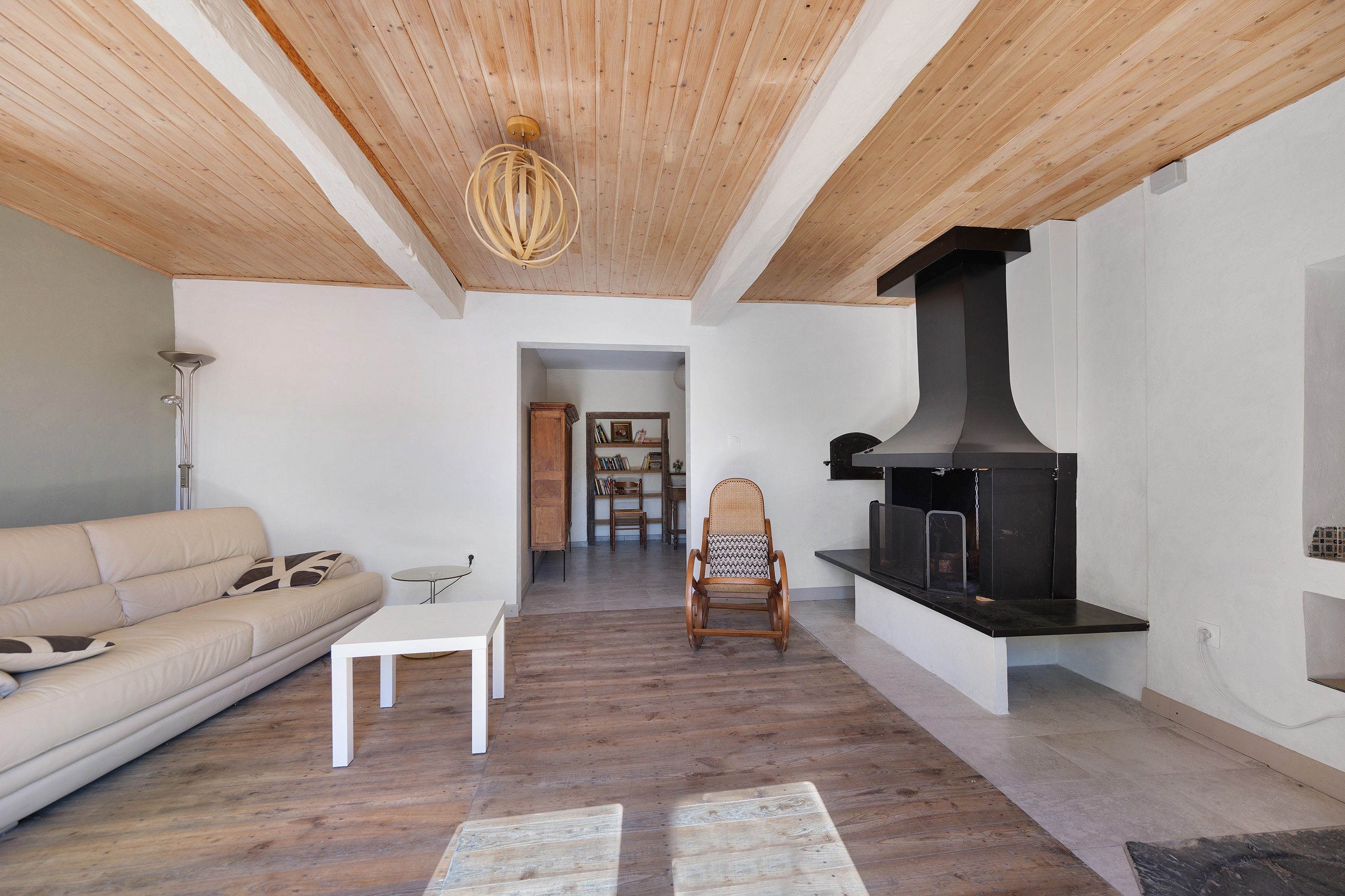A living room with a sofa, a small table, a wooden floor, a ceiling with wooden beams, and a fireplace.