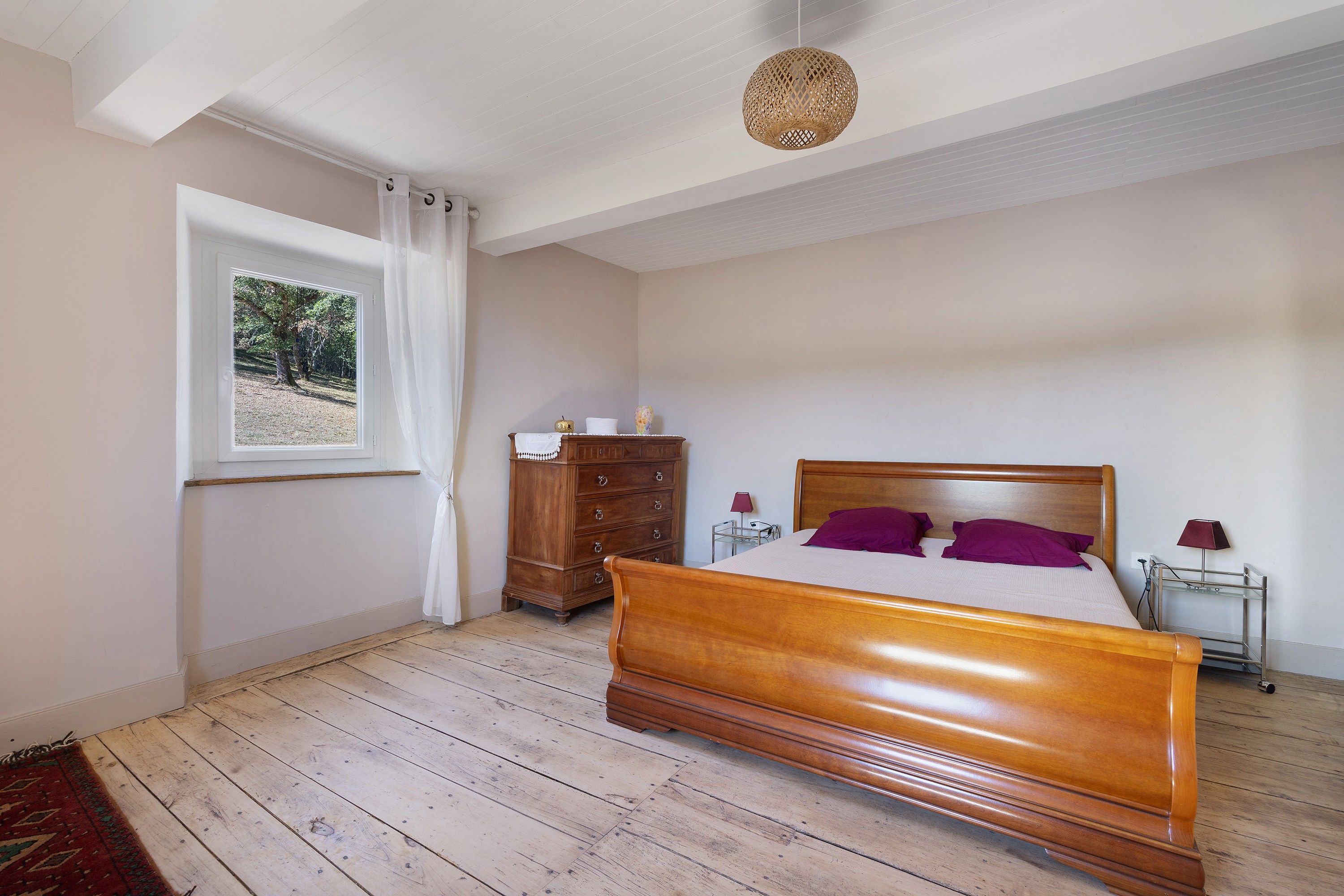 Une chambre à coucher avec un lit en bois, une commode, une table de chevet, une lampe et une fenêtre donnant sur des arbres.