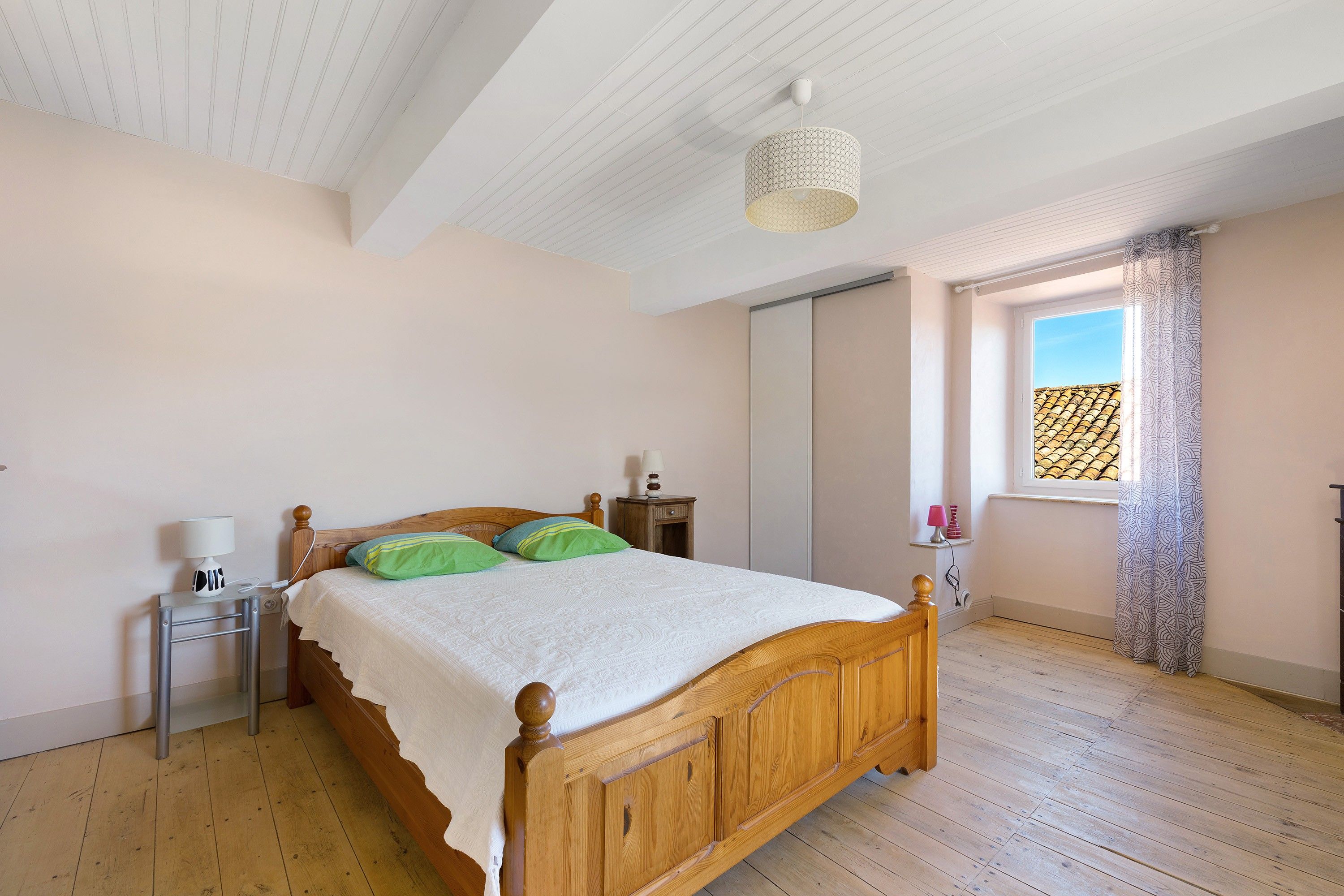Een slaapkamer met een houten bed, een nachtkastje, een lamp, een raam met gordijnen en een plafondlamp.