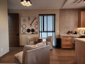 Een moderne woonkamer met een bank, een salontafel en een eethoek met tafel en stoelen. Er is een keuken met kastjes en een aanrecht. Een groot abstract schilderij hangt aan de muur, en er is een raam met blinds te zien.