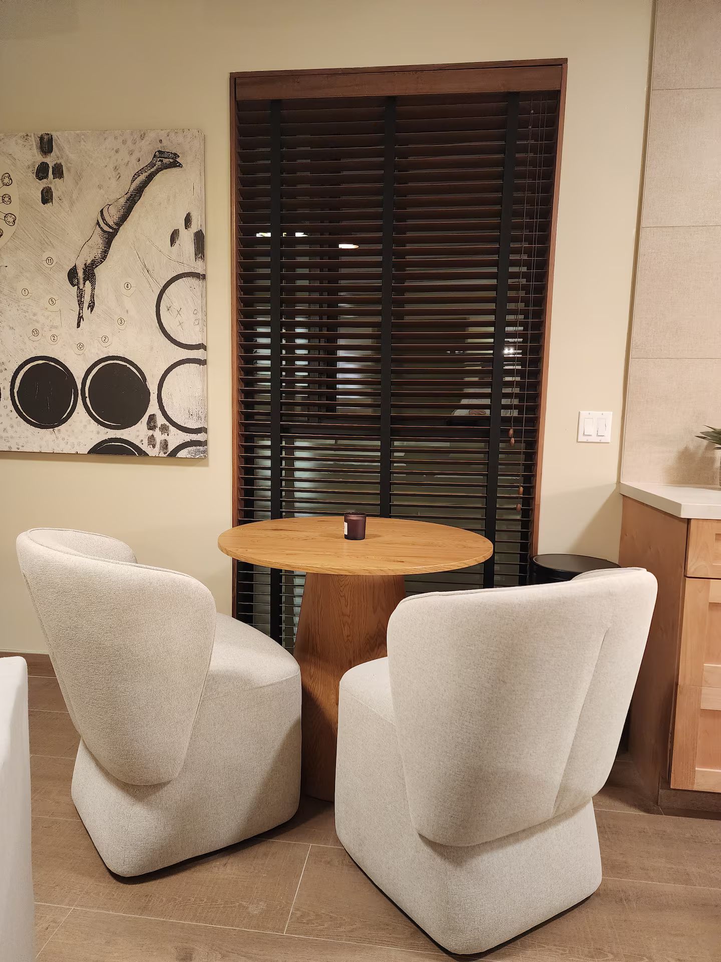 Quarto com mesa de madeira redonda, duas poltronas brancas, um quadro na parede e uma janela com persianas.