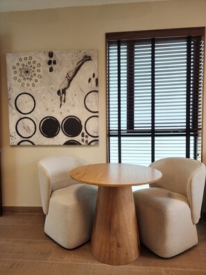 Een kamer met een ronde houten tafel, omringd door twee beige fauteuils, een groot abstract schilderij aan de muur en een raam met zonwering.