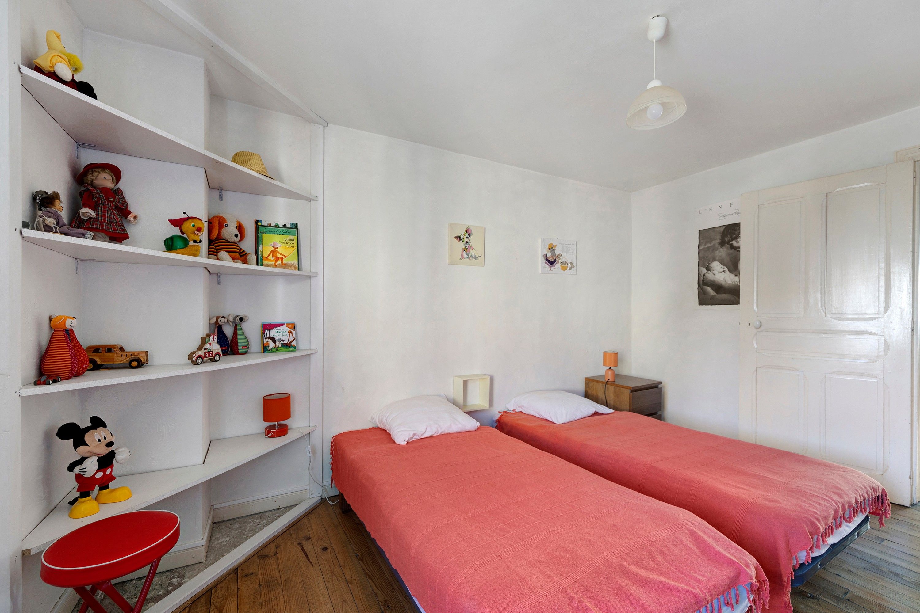 A room with two beds, a red chair, a shelf with toys and books, and a door.