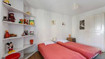 A room with two beds, a red chair, a shelf with toys and books, and a door.