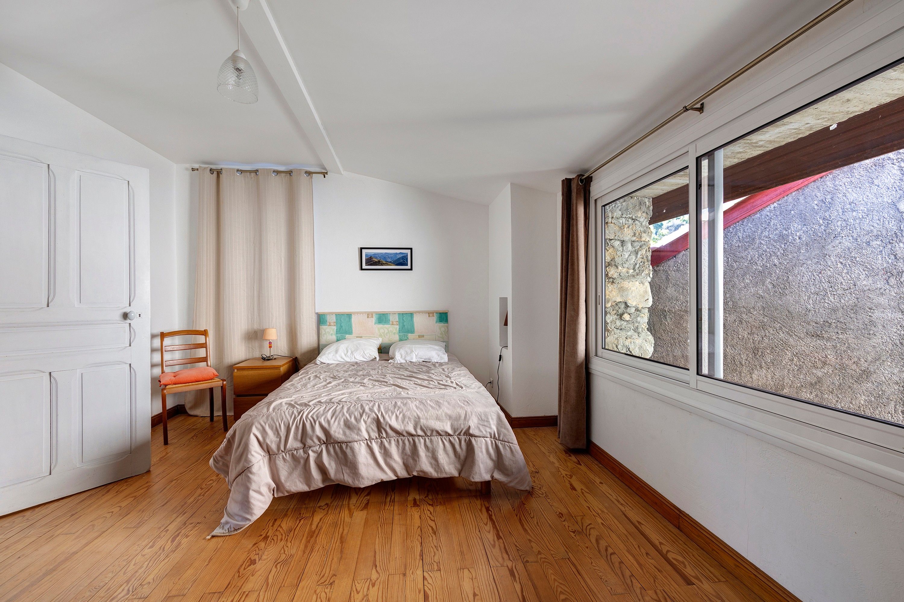 A bedroom with a bed, a chair, a nightstand, a window with curtains, and a picture on the wall.