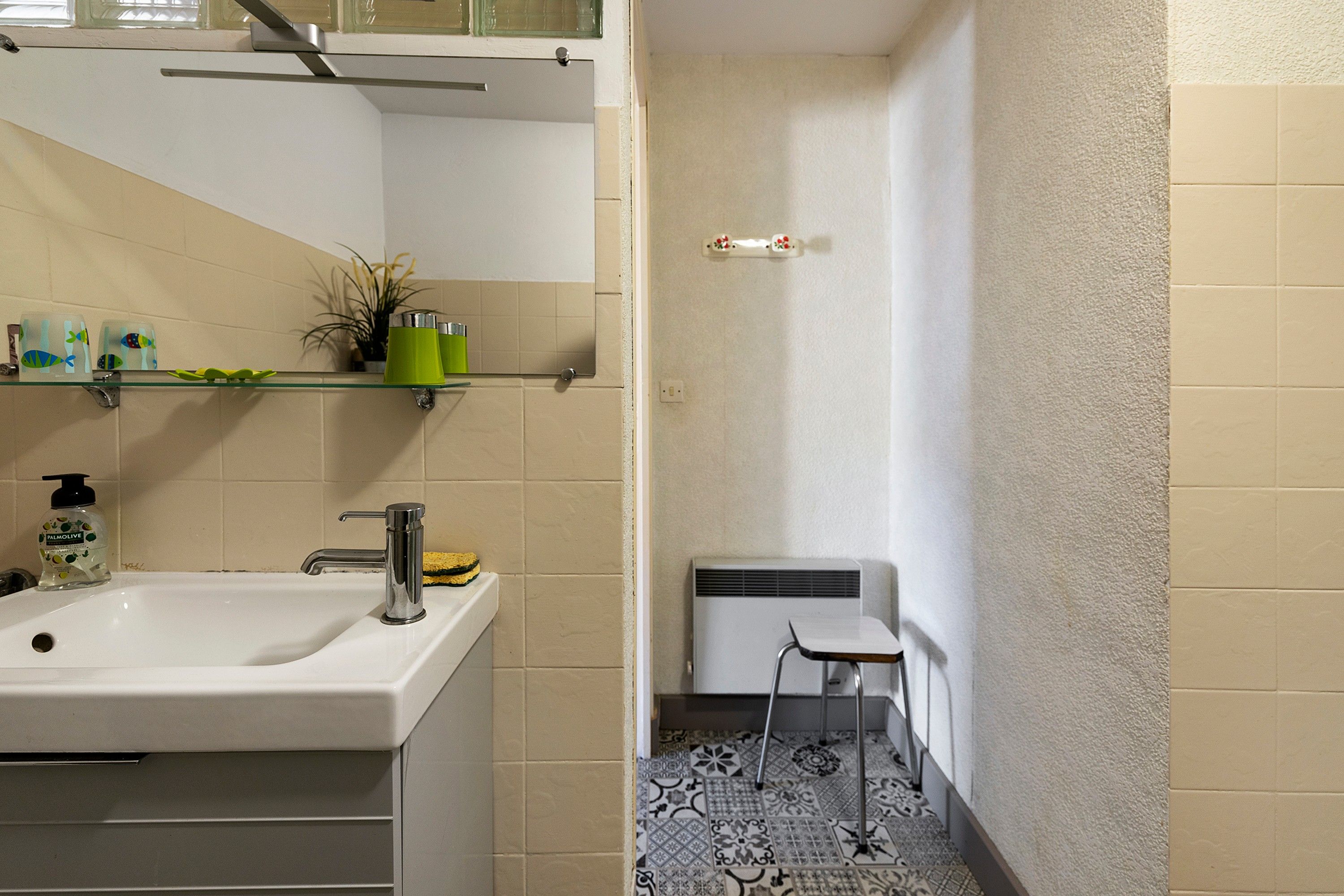 Ein Badezimmer mit Waschbecken, Spiegel, Ablagen für Toilettenartikel, Heizkörper und Fliesenboden mit Muster.