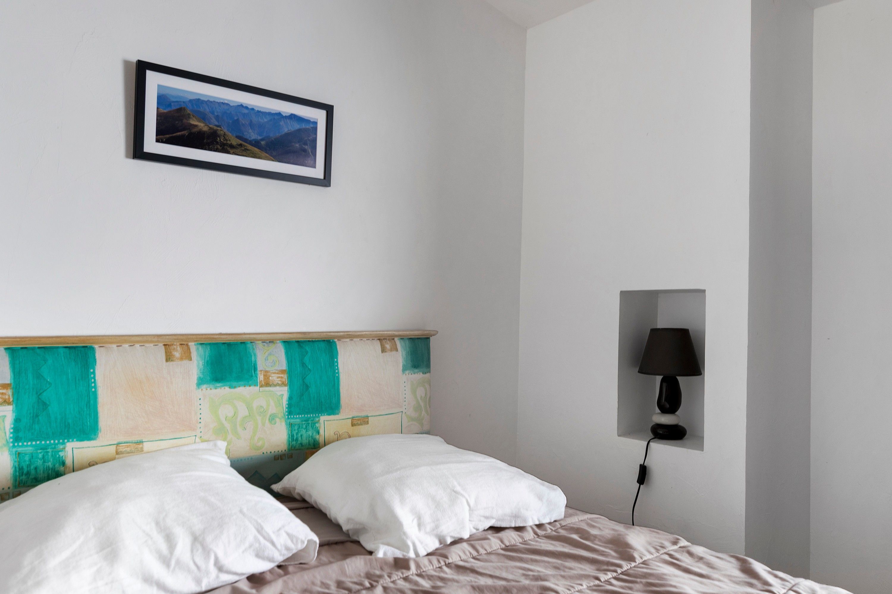 A bedroom with a bed, white pillows, a headboard with a colorful panel, a framed picture on the wall, and a bedside lamp.