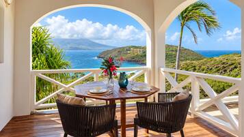 A wooden deck with a table and chairs, overlooking a coastal landscape with a view of the ocean and hills.