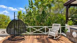Una terraza de madera con una silla de mimbre y una mecedora, rodeada de abundante vegetación y una cerca blanca.