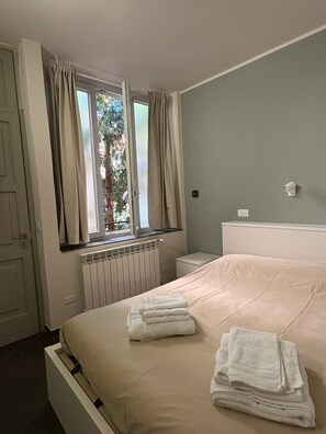 Quarto com cama, janela com cortinas, radiador e mesa de cabeceira.