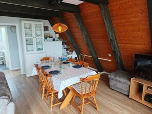 A dining area with a table set for six, surrounded by wooden chairs, in a room with exposed wooden beams and a television stand.