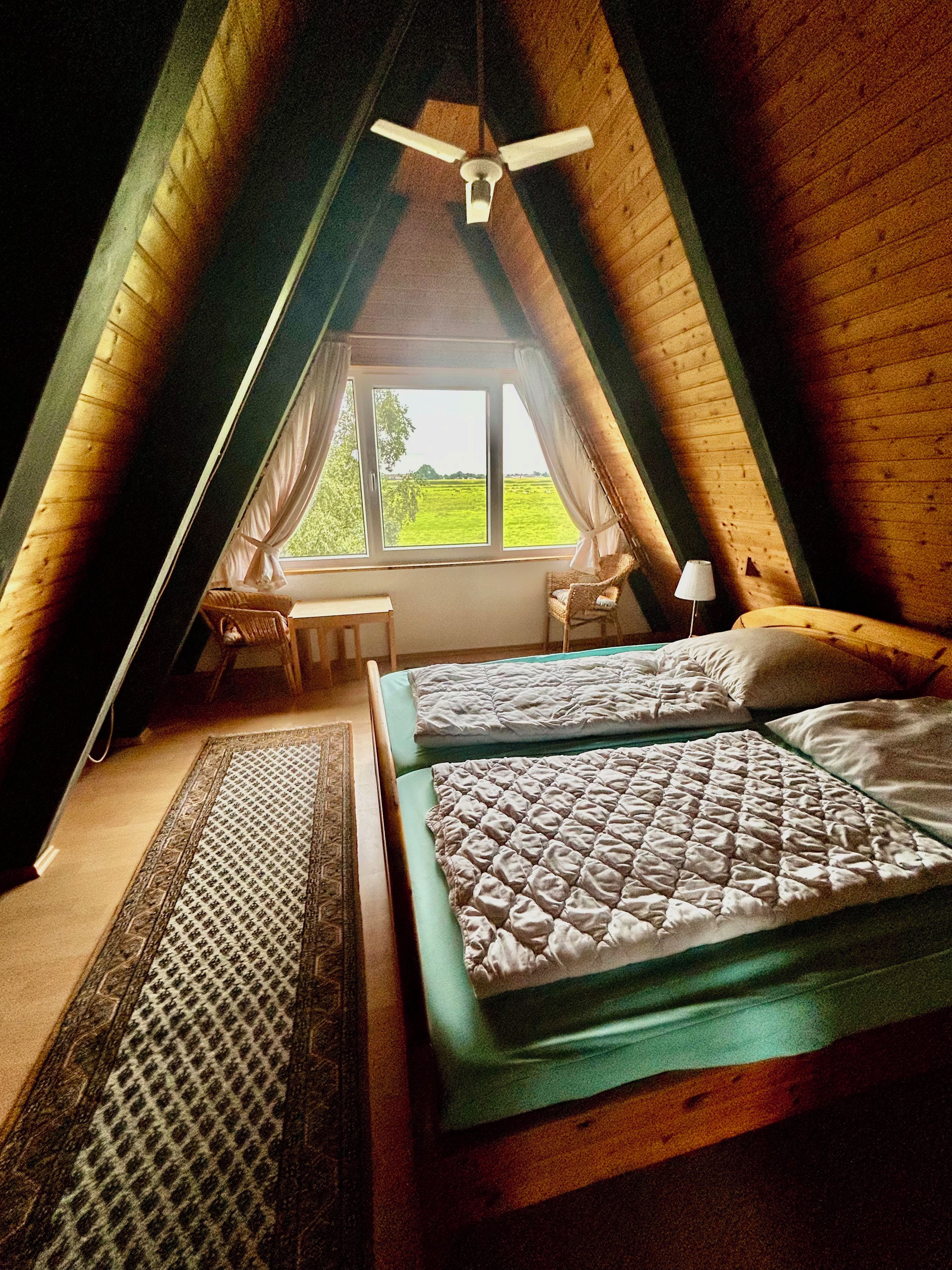 Une pièce avec un plafond en pente, un lit avec un matelas vert, une table de chevet, une lampe et une vue sur l’extérieur par une fenêtre.