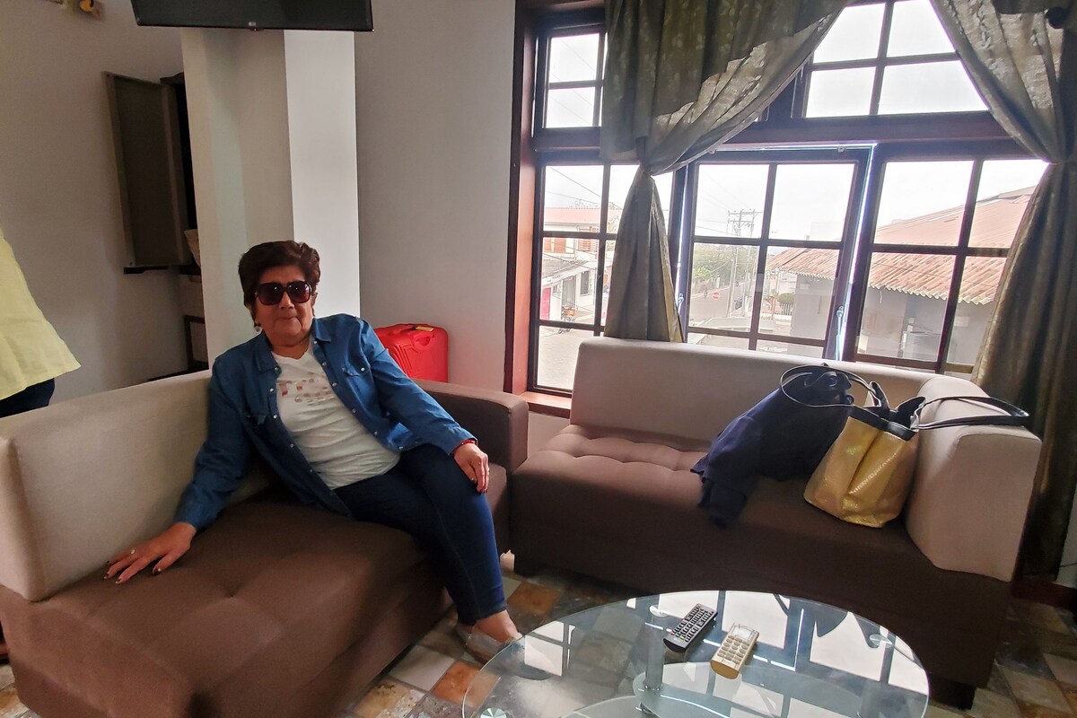 A living room with a brown sofa, a glass coffee table, and a view through a window with curtains.
