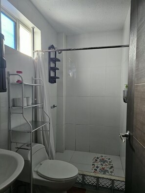 A small bathroom with a toilet, a sink, a shower with a curtain, and a shelf with toiletries. - CASA RENTERA D'ELIAS (Puerto Baquerizo Moreno)