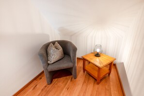 Une pièce d’angle avec un plancher en bois, un fauteuil gris avec un coussin et une petite table en bois sur laquelle se trouve une lampe.