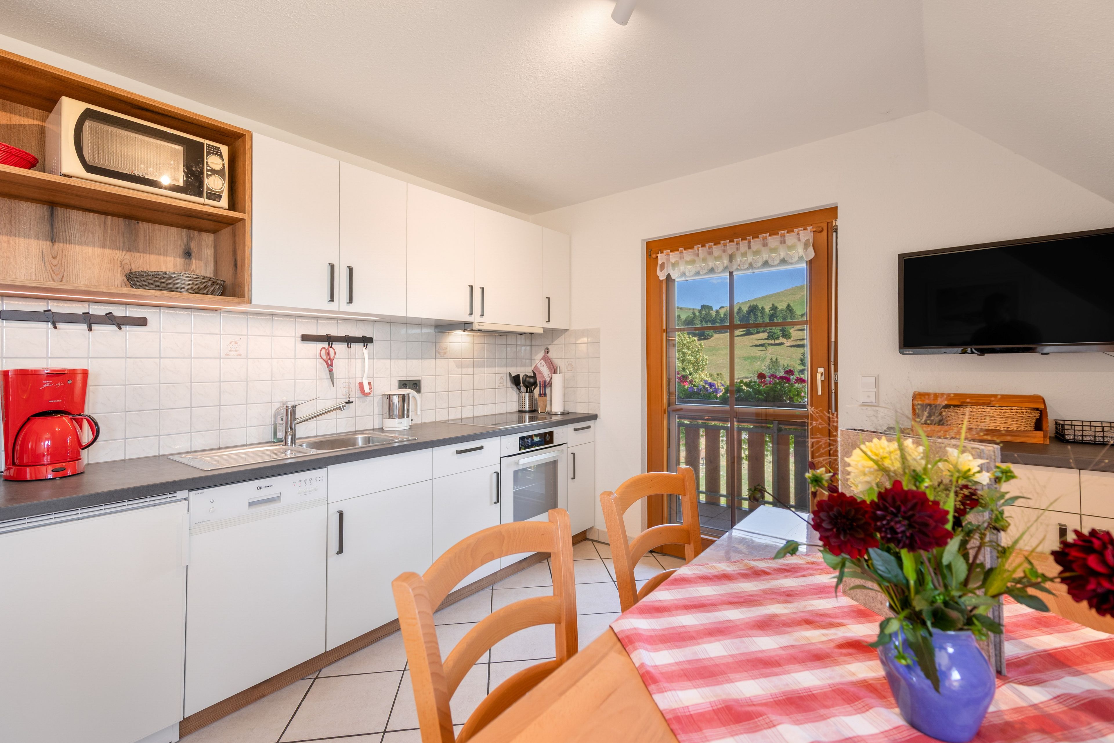 Une cuisine moderne avec des meubles blancs, une machine à café rouge et un coin repas agrémenté d’une nappe à carreaux et de chaises.