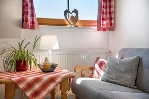 Ein gemütliches Wohnzimmer mit einer Couch, einem Tisch mit rotem und weißem Karomuster, einer Topfpflanze, einer Lampe und einer herzförmigen Dekoration am Fensterbrett.