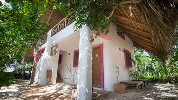 A two-story house with a thatched roof, white walls, and pink accents. There are potted plants and a wooden bench outside.