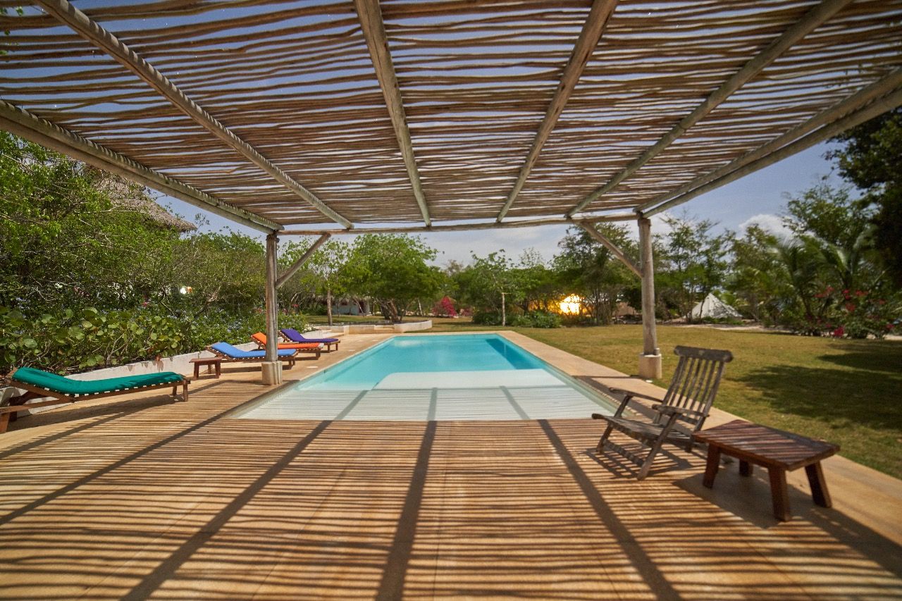 An outdoor swimming pool area with a wooden deck, lounge chairs, and a pergola providing shade.