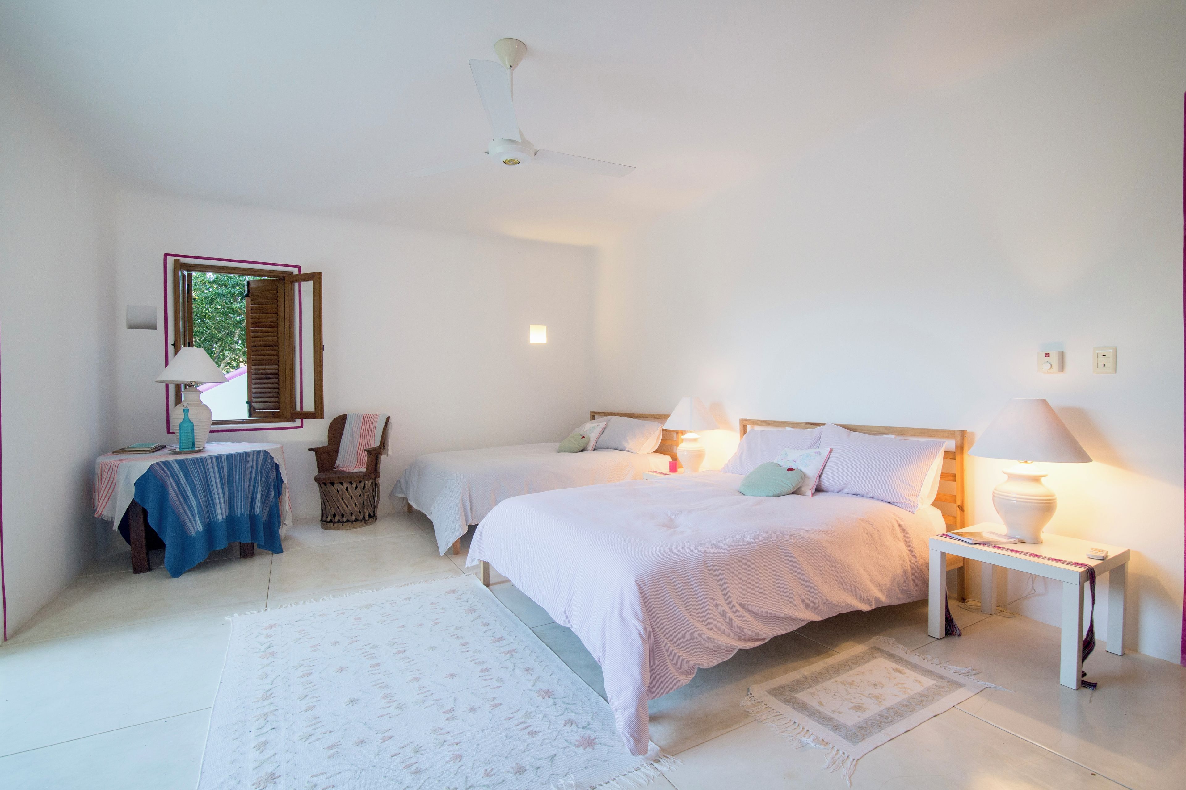 A bedroom with two single beds, a bedside table with a lamp, a small table with a lamp, a chair, and a window with shutters.
