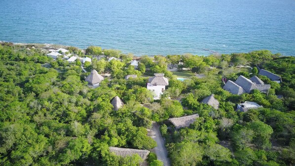 An aerial view of a resort with thatched-roof cottages surrounded by dense greenery and a clear view of the sea.