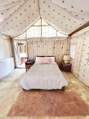 A tented room with a bed, a nightstand, a lamp, and a small table with books and a bowl. - Ki-Ra Holistic Living (San Rafael del Yuma)