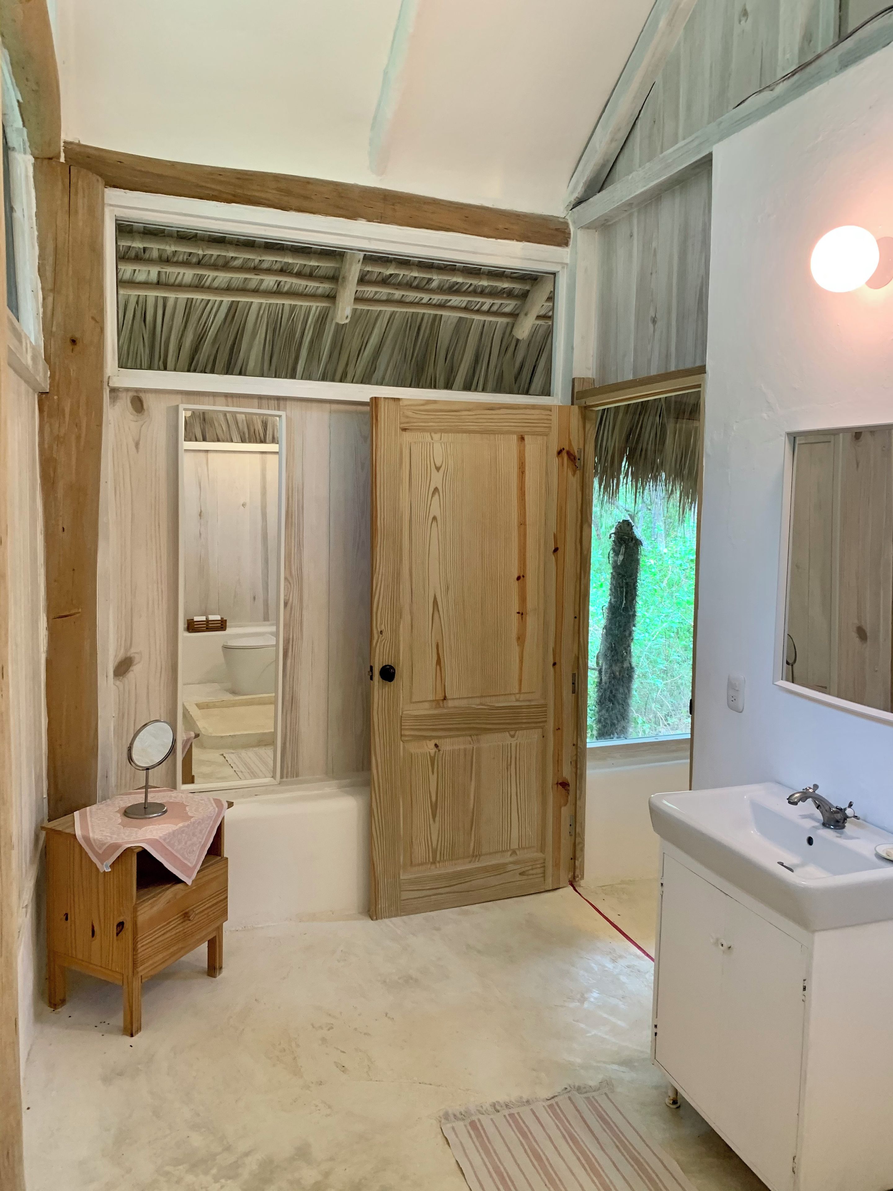 A bathroom with a wooden door, a small vanity with a mirror, a white sink, and a window with a view of greenery.