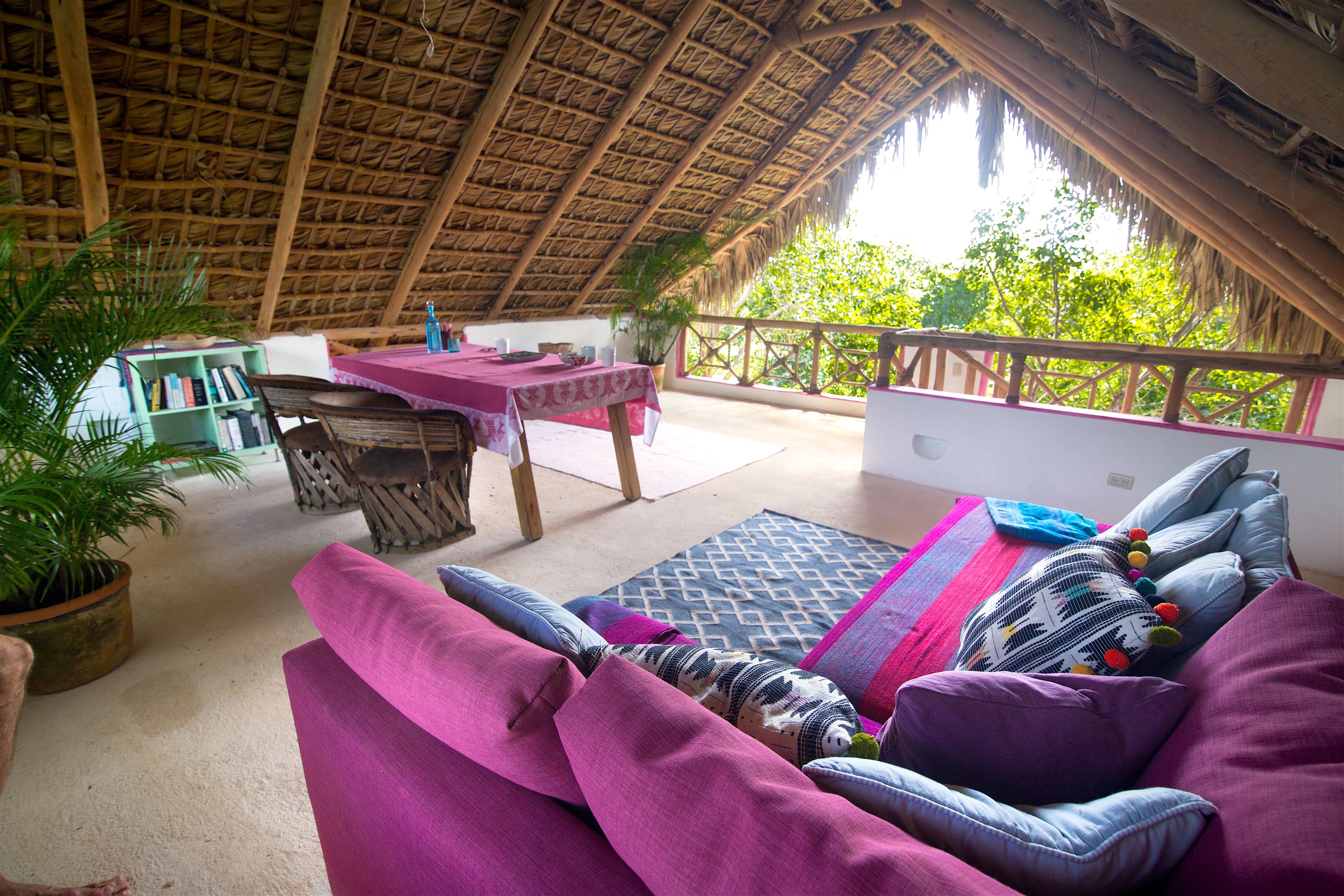 A room with a thatched roof, a sofa with cushions, a table with a pink tablecloth, and a view of greenery outside.